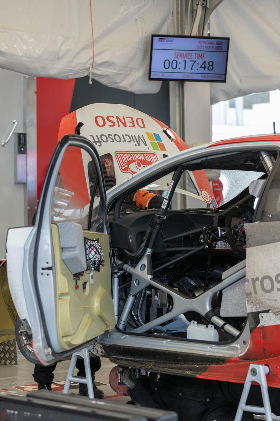 Zwei Mechaniker reparieren einen Rallyewagen in einer Boxengasse.